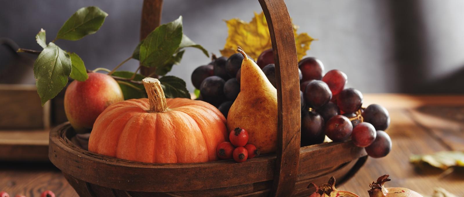 Wooden basket filled with autumn harvest including pumpkin, pear, apple, grapes and berries with autumn leaves.