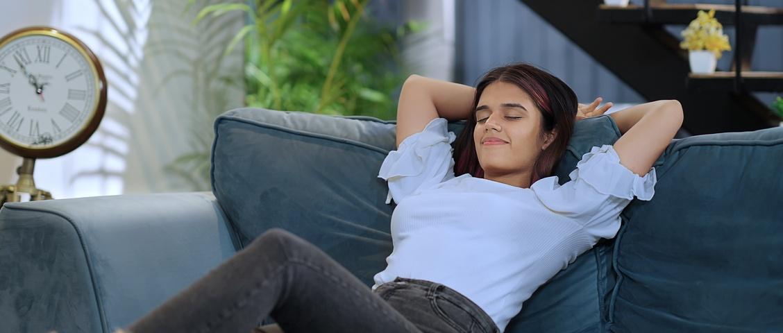 Woman relaxing on blue sofa with eyes closed, wearing white top and jeans, with vintage clock and plants nearby.