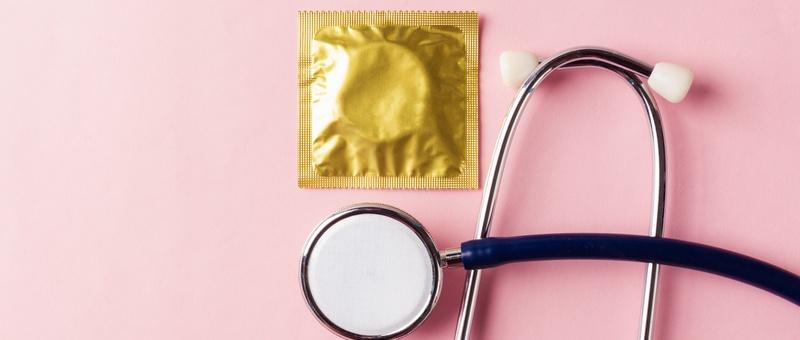 Condom in wrapper beside stethoscope against pink background.