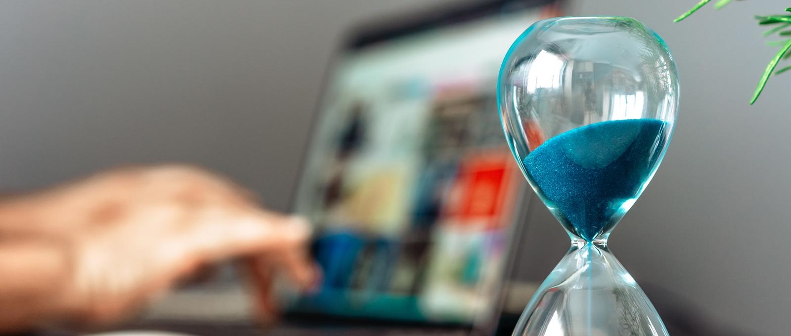 Glass hourglass with blue sand next to a laptop being used, symbolising time management in digital work.