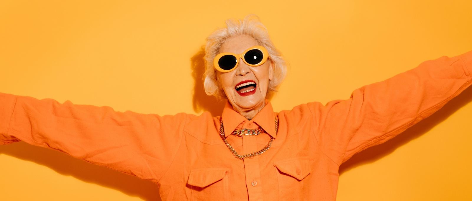 Smiling senior stylish woman wearing trendy sunglasses spreads hands, wants to hug someone, glad to see you isolated on yellow background.