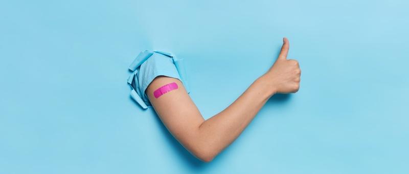 Female arm in blue t-shirt with purple plaster in thumbs-up position bursting through blue paper background.