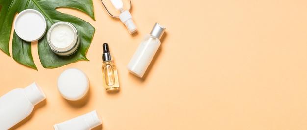 Variety of white and clear creams and serums in assorted containers with dark green leaf detail against a beige background.