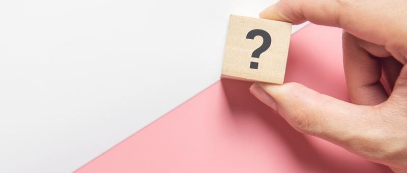 Hand holding question mark printed on wooden cube against diagonally split white and pink background.