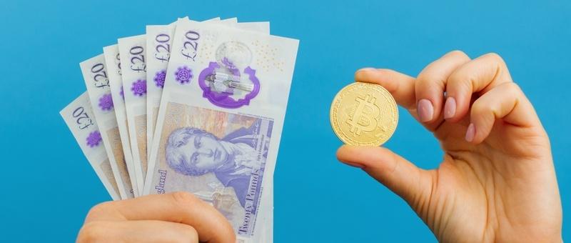 Hands holding fan of British £20 notes and one gold Bitcoin coin against blue background.