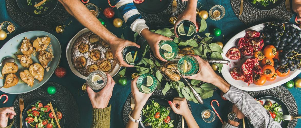 Hands toasting drinks over festive table with apple cake, pastries, fresh fruits and holiday decorations.