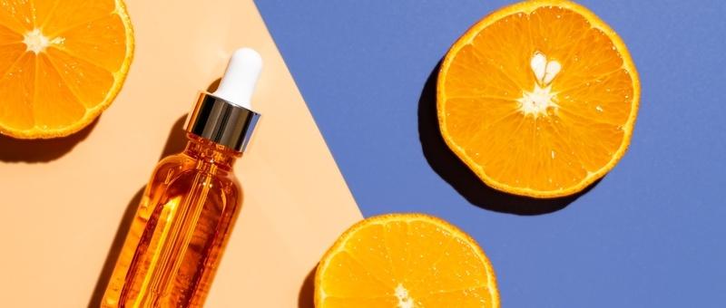 Orange serum bottle with dropper alongside fresh orange halves on peach and blue background.