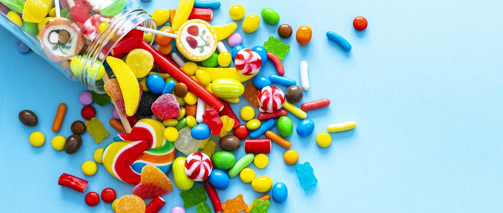 Large group of all sort of multicolored sweets spilling out from a glass jar shot from above on blue background.