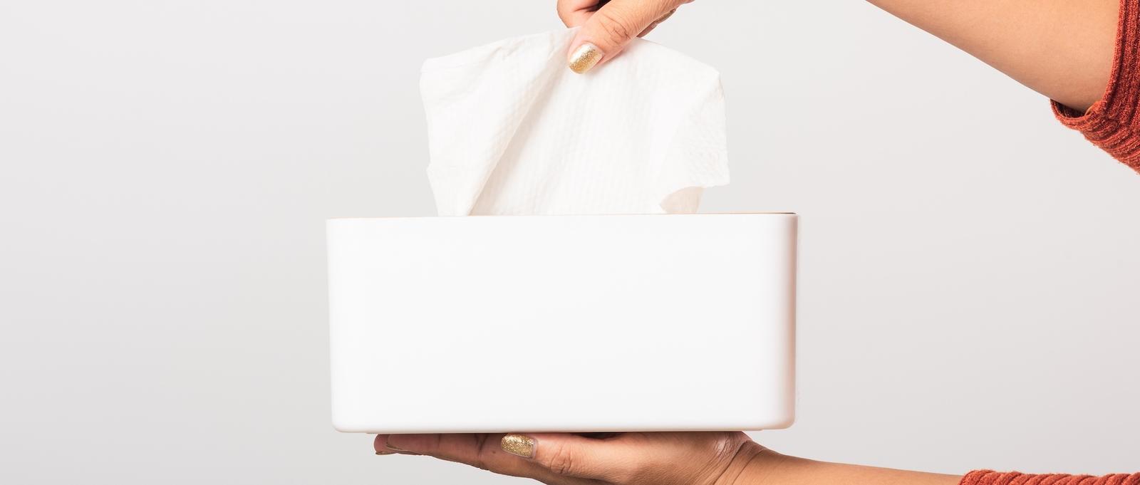Hand with gold nail polish pulling a tissue from a white rectangular box against a light grey background.