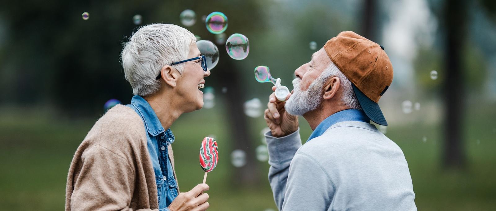 Senior couple enjoying playful moment outdoors with bubbles and a lollipop, sharing laughter in a park setting.