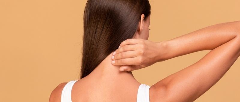 Back view of woman with long brown hair in white vest, holding neck against tan background.