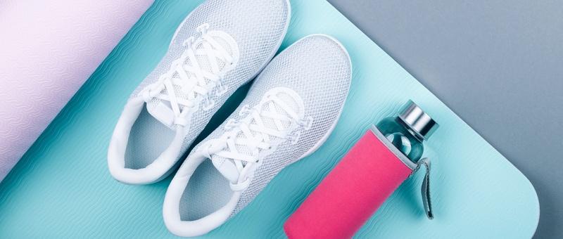Top view of white sport shoes beside pink metal drinks bottle resting on pink and blue yoga mat against pale grey background.