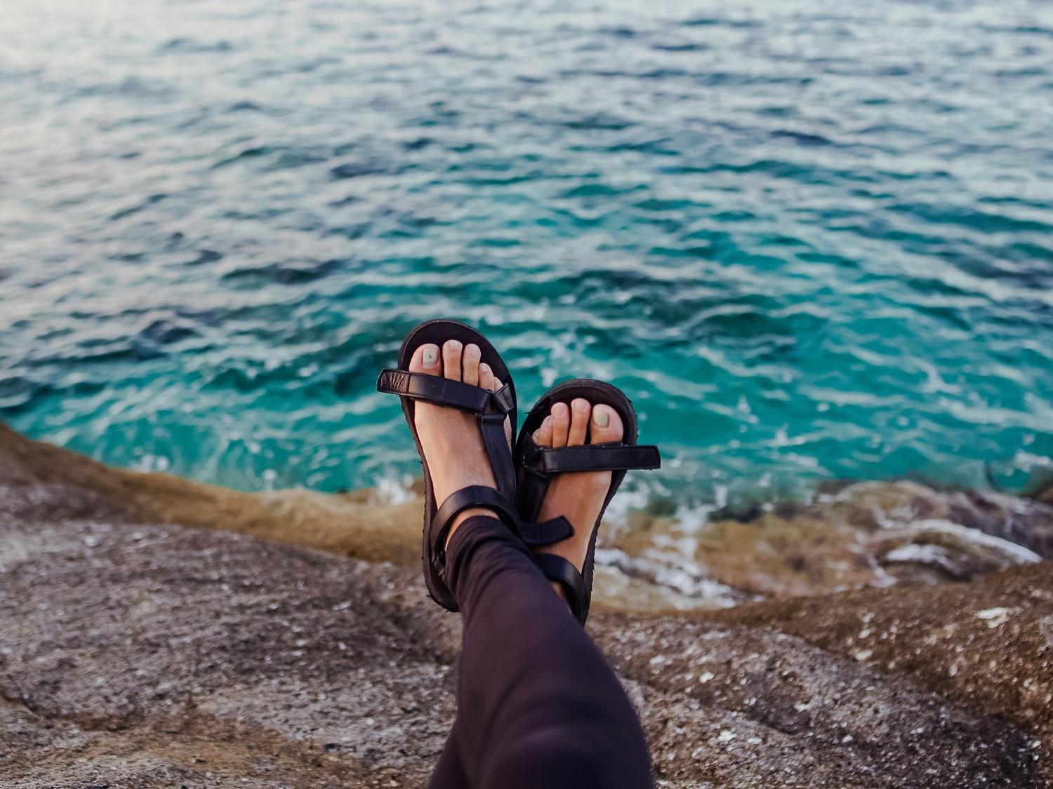 Feet in black sandals dangling over turquoise ocean water, viewed from a rocky coastal edge.