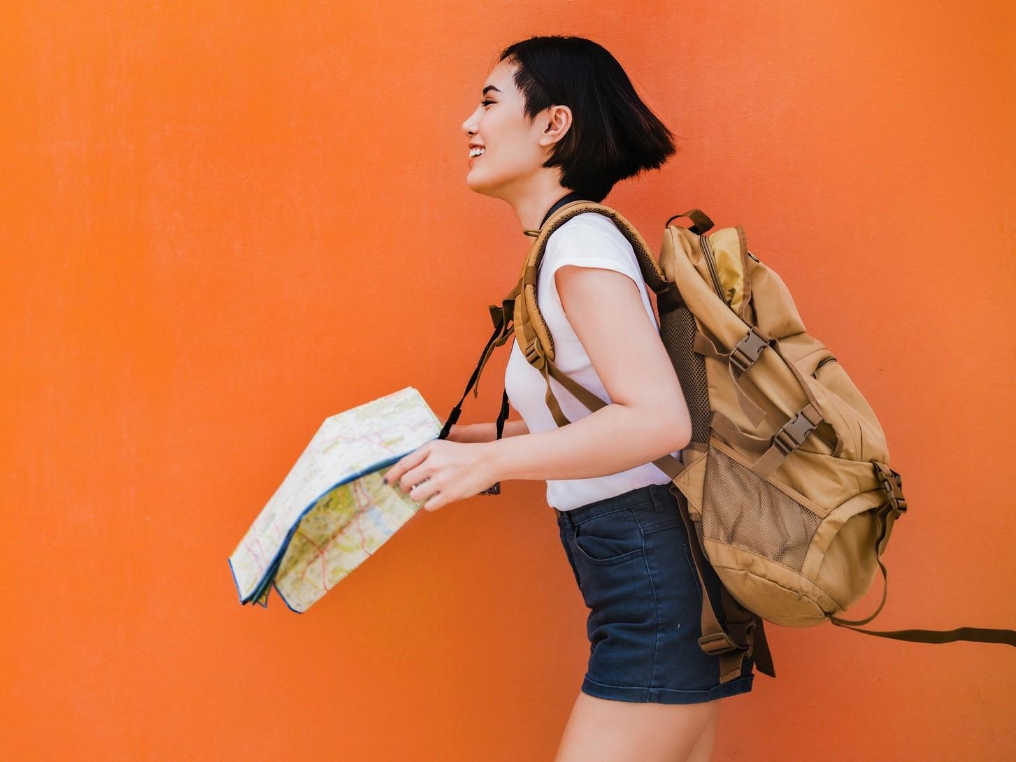 Person with short dark hair wearing a backpack and holding a map, smiling against a bright orange background.