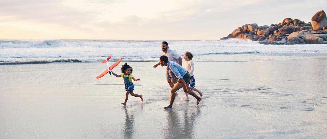 Family running along shoreline at sunset, child holding red toy plane, rocky coastline in background.