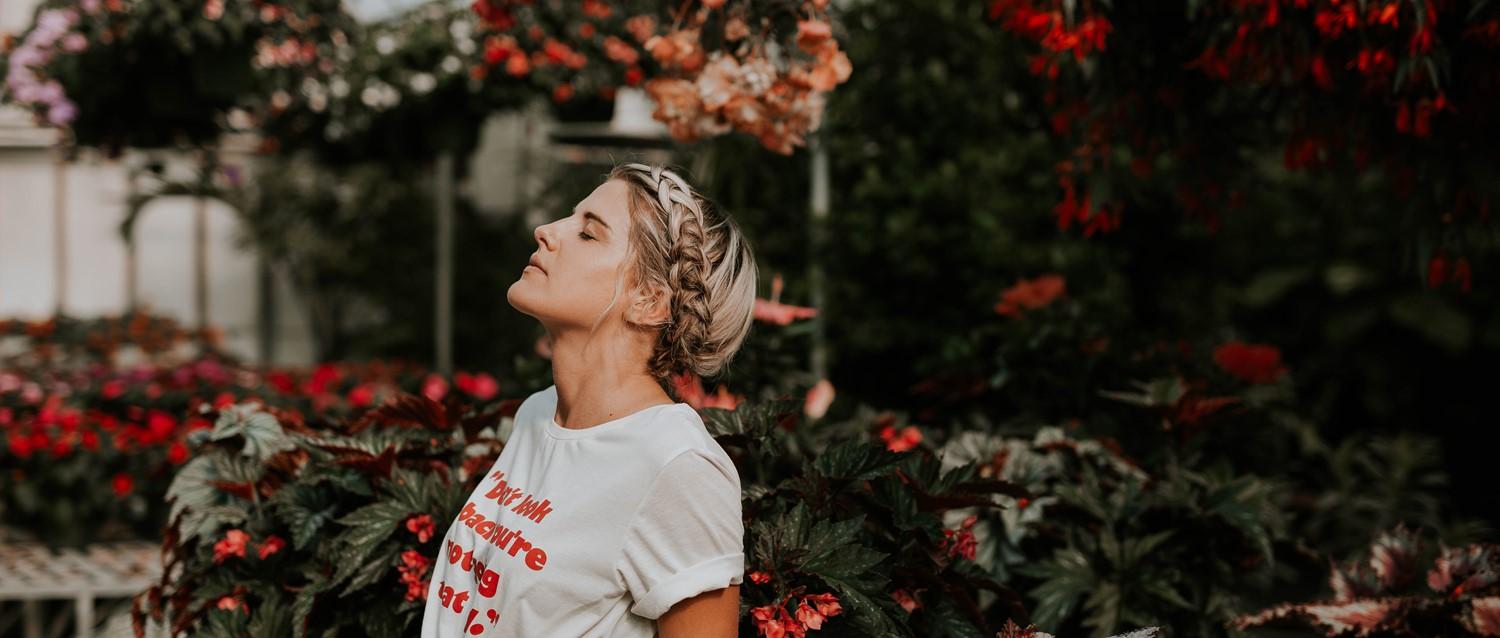 Female with braided blonde hair in white t-shirt surrounded by vibrant red flowers and lush greenery in a garden.