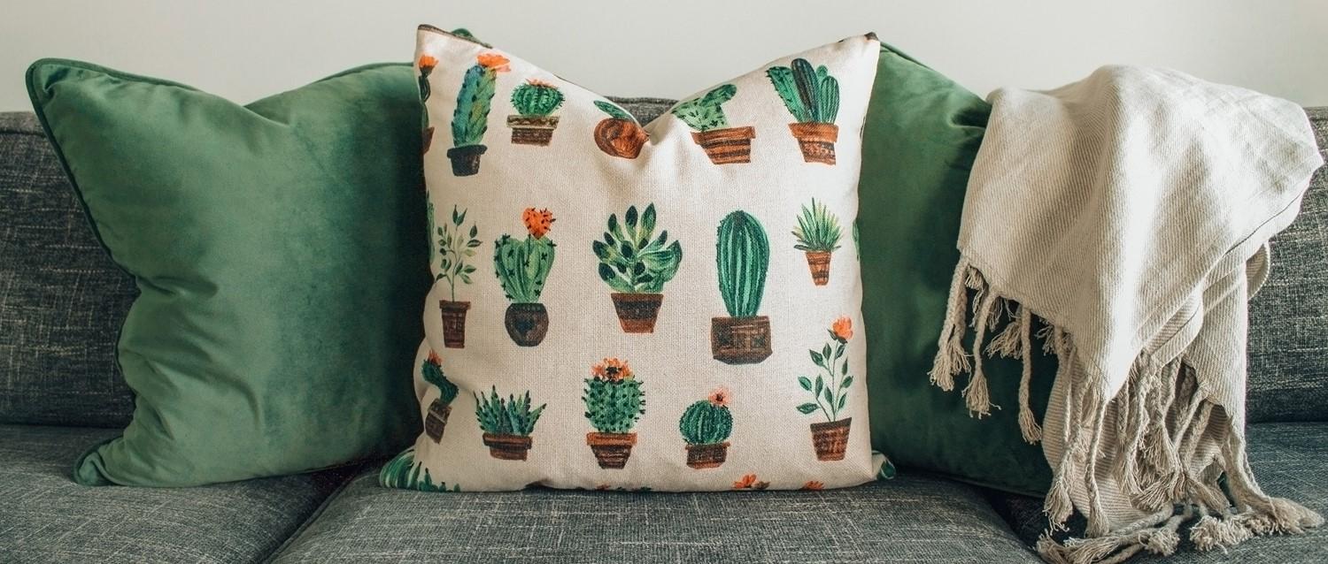 Decorative cushions on grey sofa: green velvet pillows, cream cushion with cactus pattern, and fringed throw blanket.