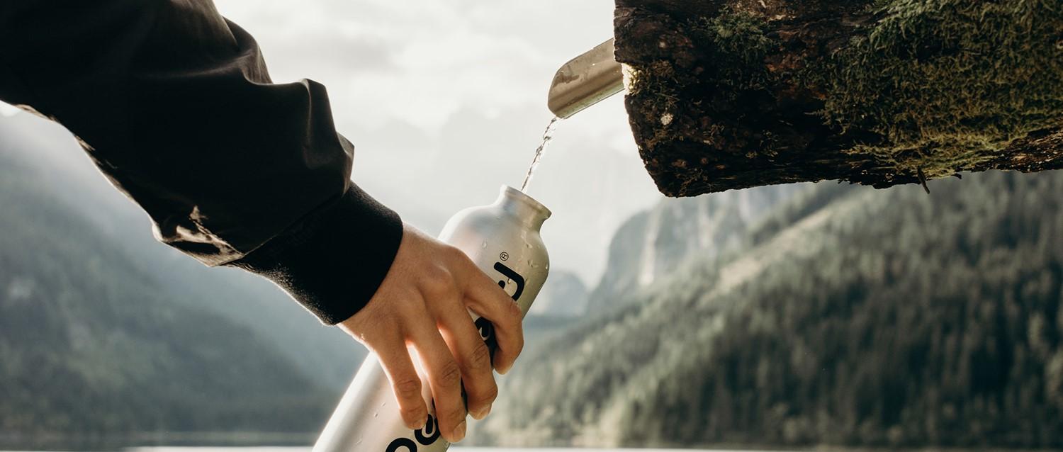 Person filling reusable water bottle from natural spring in mountainous landscape with dramatic rocky peaks in background.