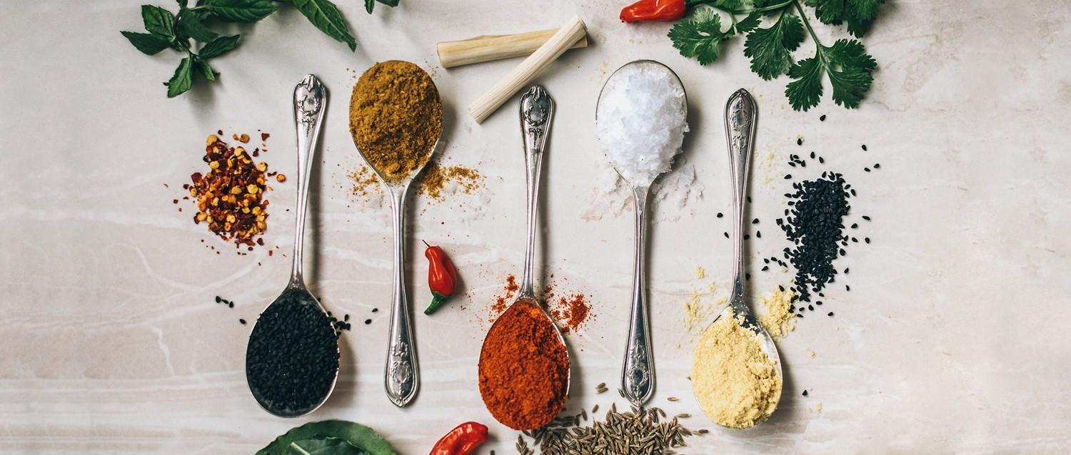 Silver spoons with colourful spices including curry, paprika, salt and black seeds, surrounded by fresh herbs and chillies.