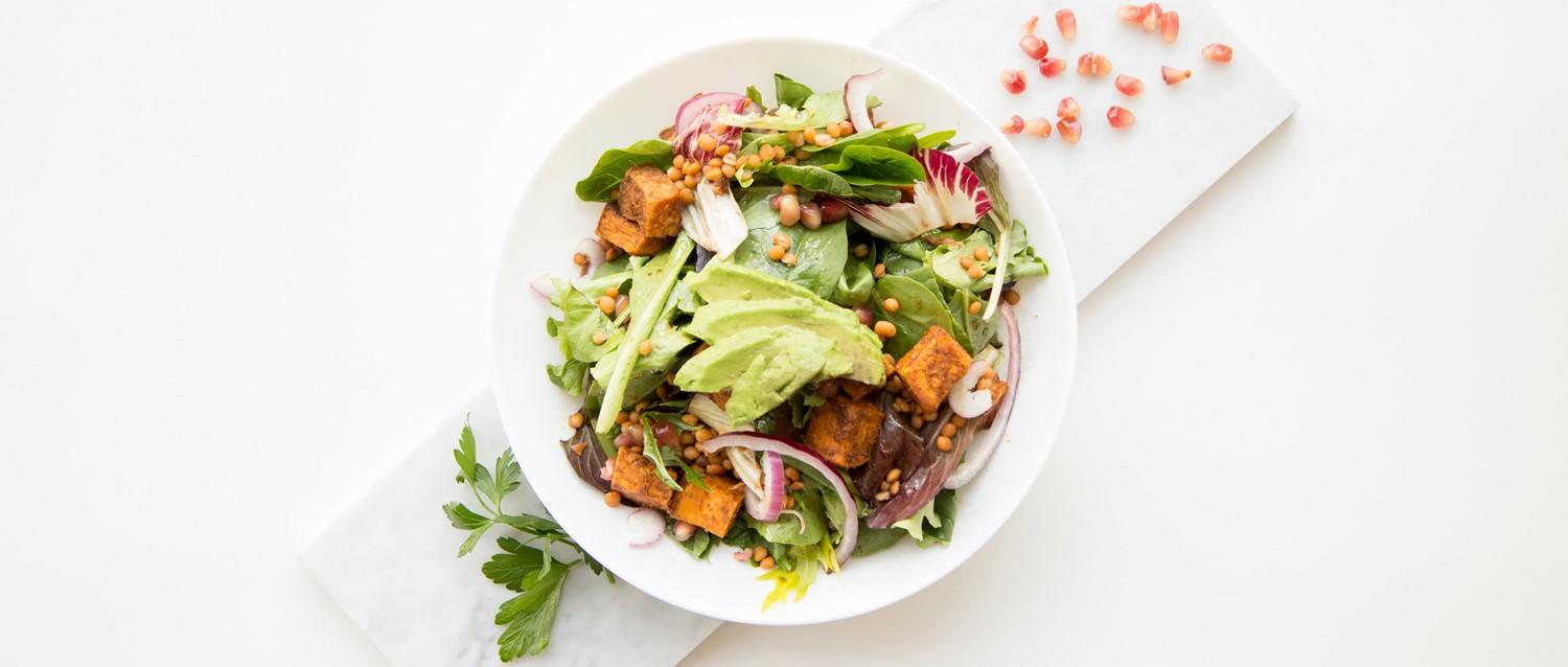 Fresh salad bowl with avocado slices, tofu cubes, lentils, mixed greens and red onions on white marble surface.