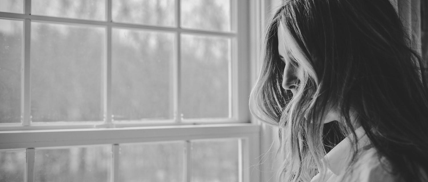 Person with long hair in profile view looking downward by a large window, captured in black and white.