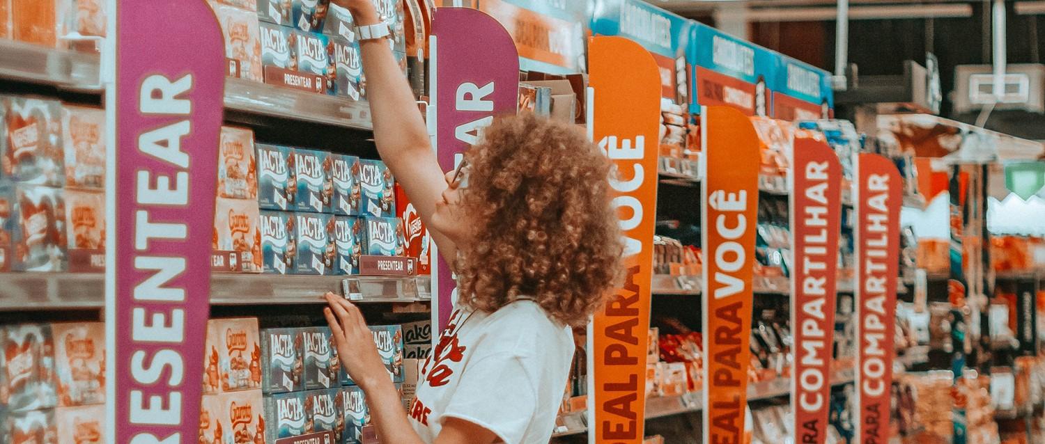Person with curly hair reaching for products in a supermarket aisle with colourful category signs in pink and orange.