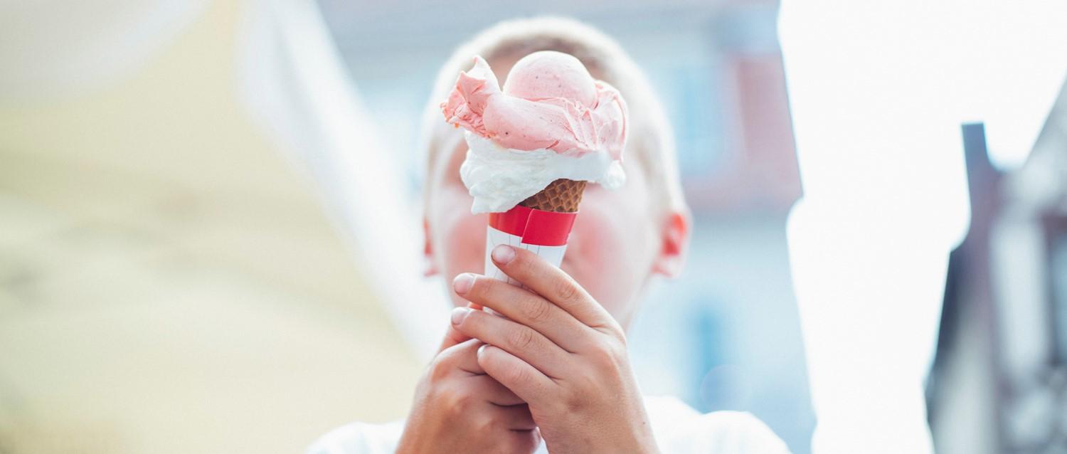 Hands holding a strawberry ice cream cone with pink scoops and whipped cream against a bright, blurred background.