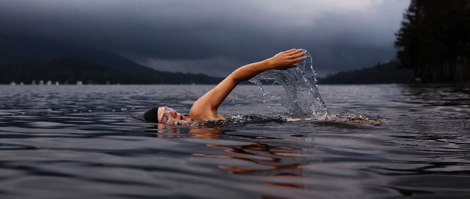 Swimmer doing front crawl in a misty mountain lake, arm creating splash against dark stormy sky backdrop.