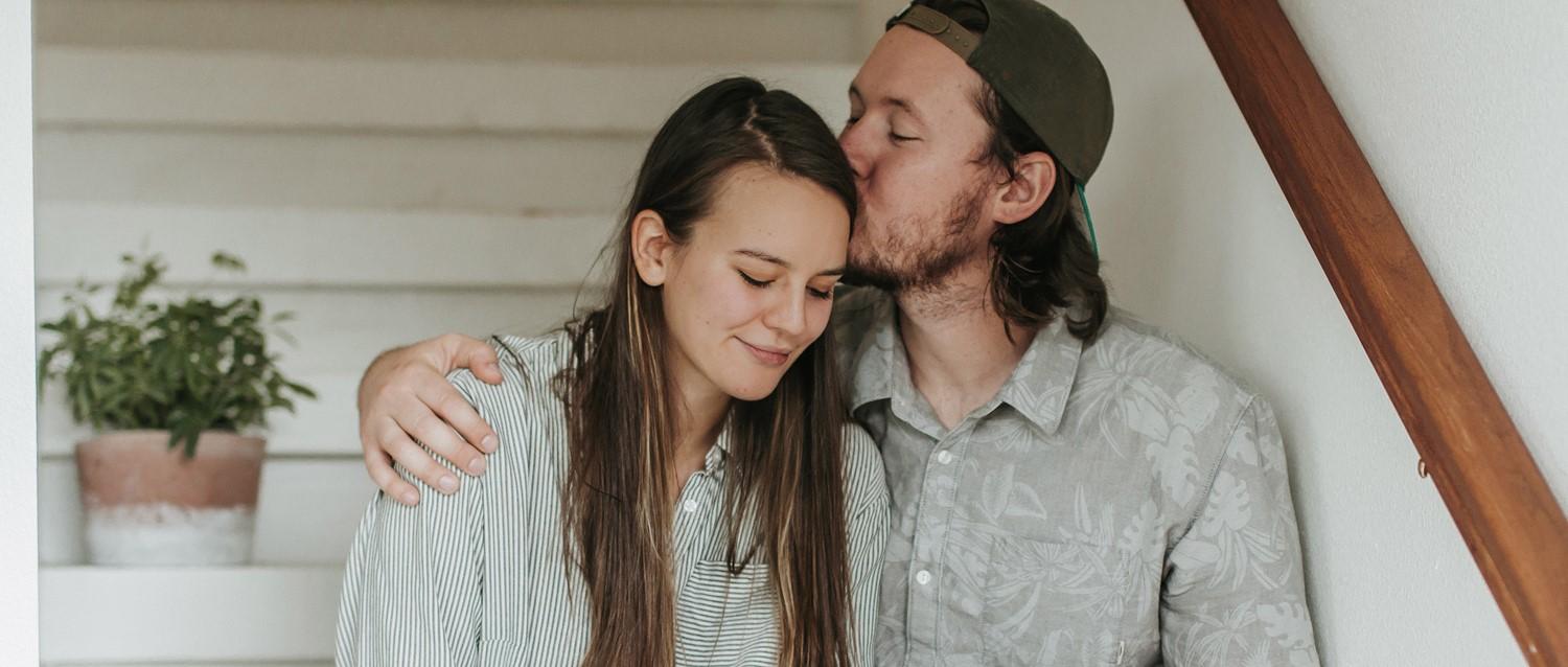 Couple sharing an intimate moment, man in cap kissing woman's forehead, both in light shirts, potted plant in background.
