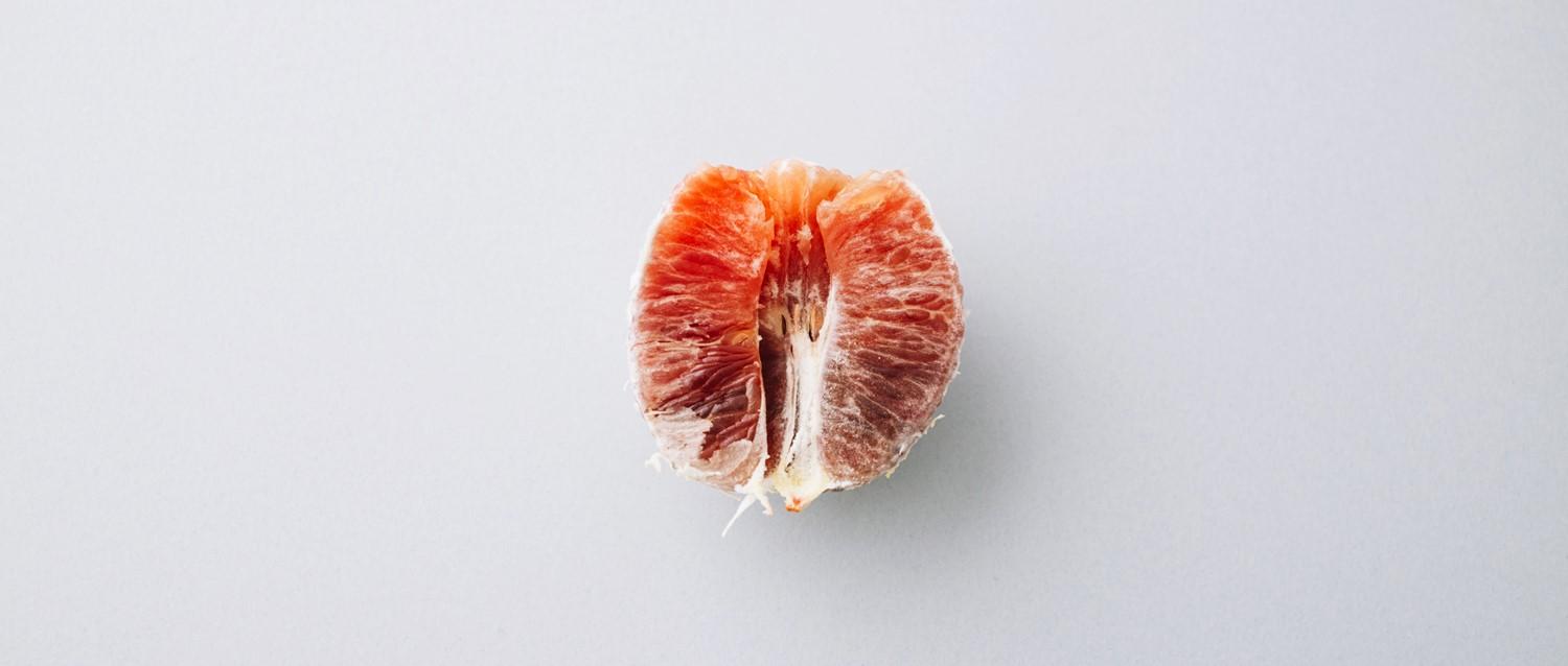 A peeled grapefruit segment showing juicy pink-red flesh against a plain light grey background.
