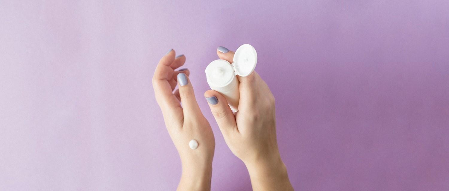 Hands with pale blue nail polish holding a white skincare container with cream on one hand against a purple background.