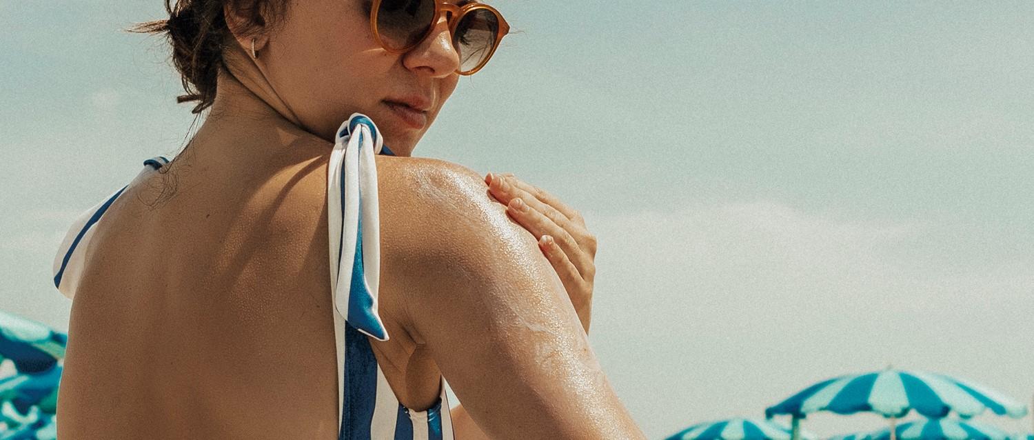 Person in round sunglasses applying sunscreen to shoulder at beach, wearing blue and white striped swimwear.