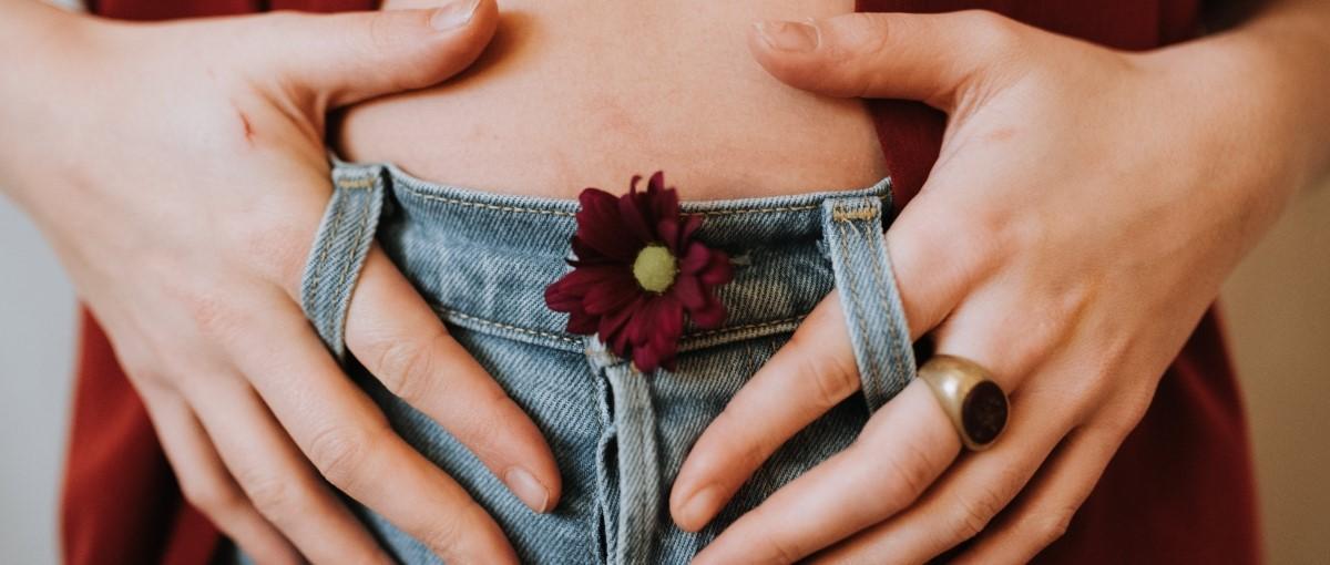 Hands forming heart shape over denim waistband with a small burgundy flower tucked in, person wearing a ring.