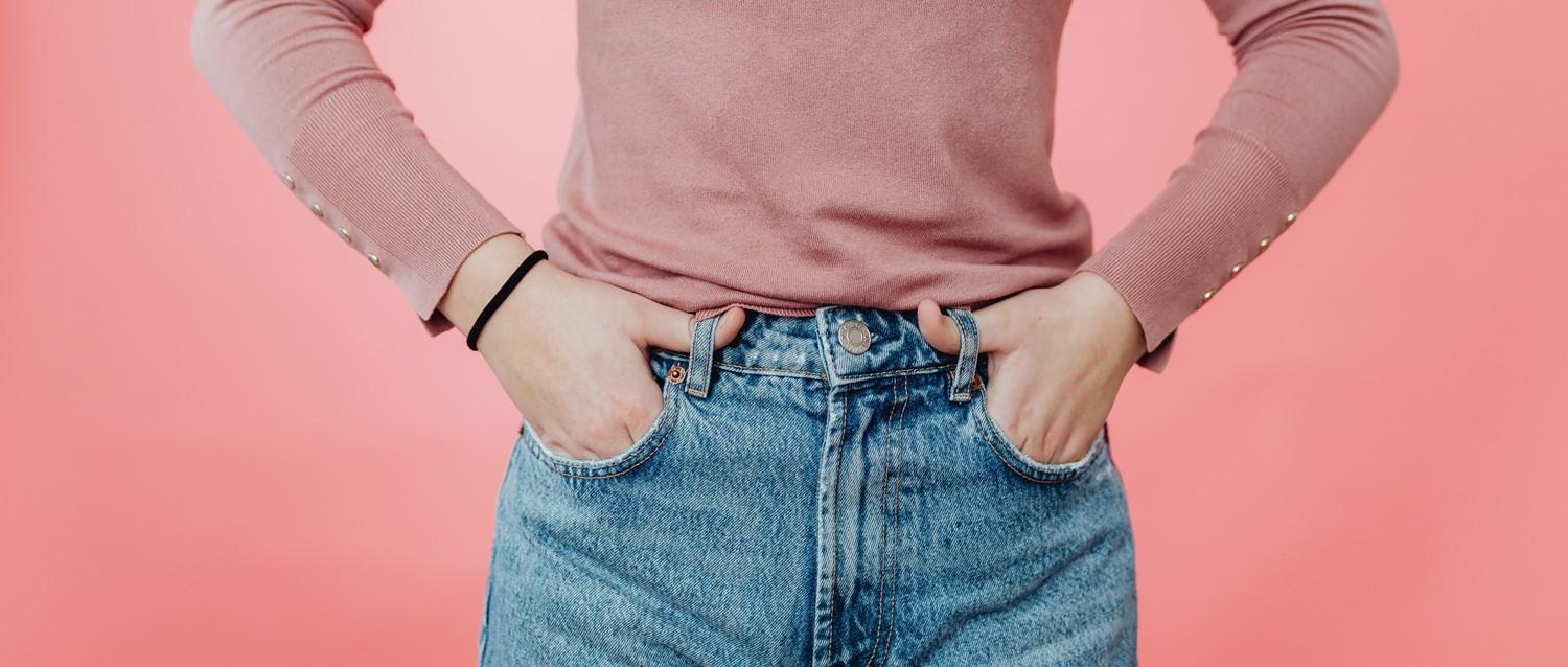 Person wearing pink jumper with hands in blue denim jeans pockets against coral pink background.