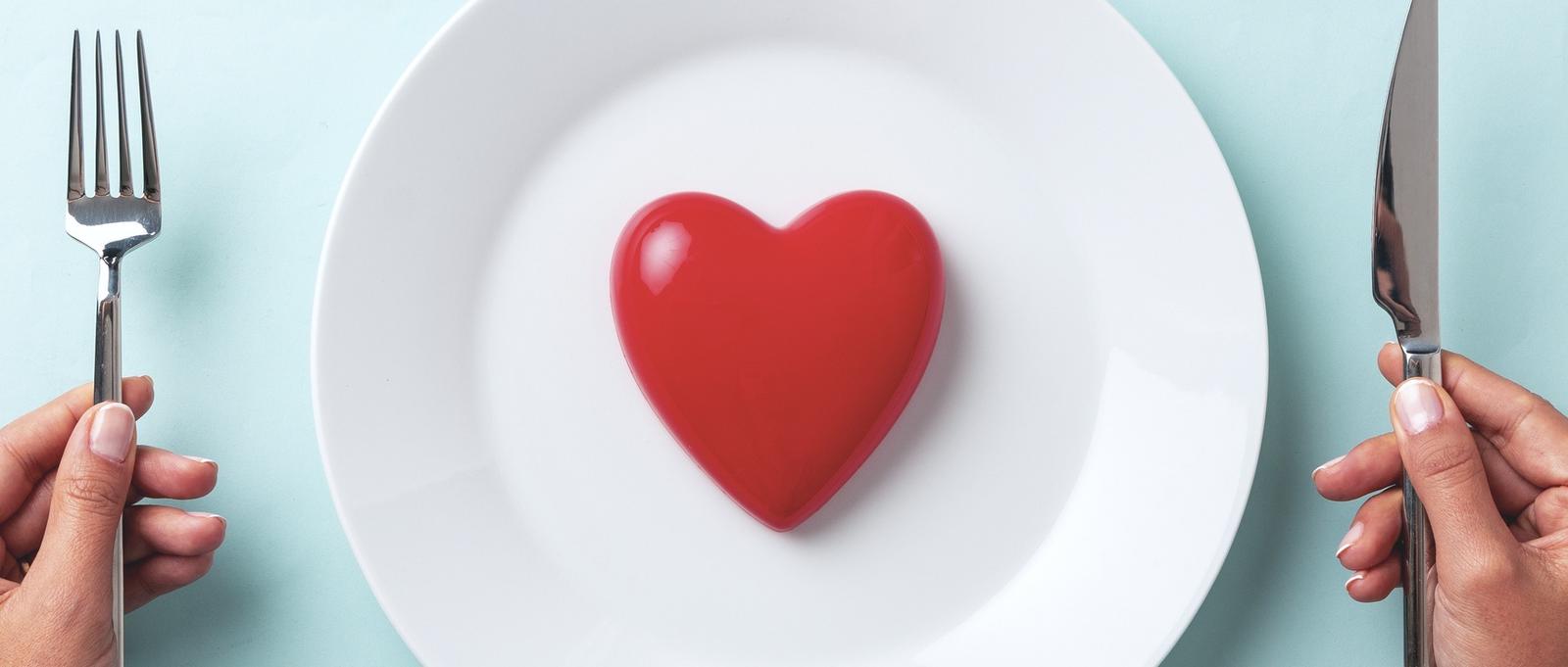 Red heart on white dinner plate against blue background with hands holding knife and fork either side